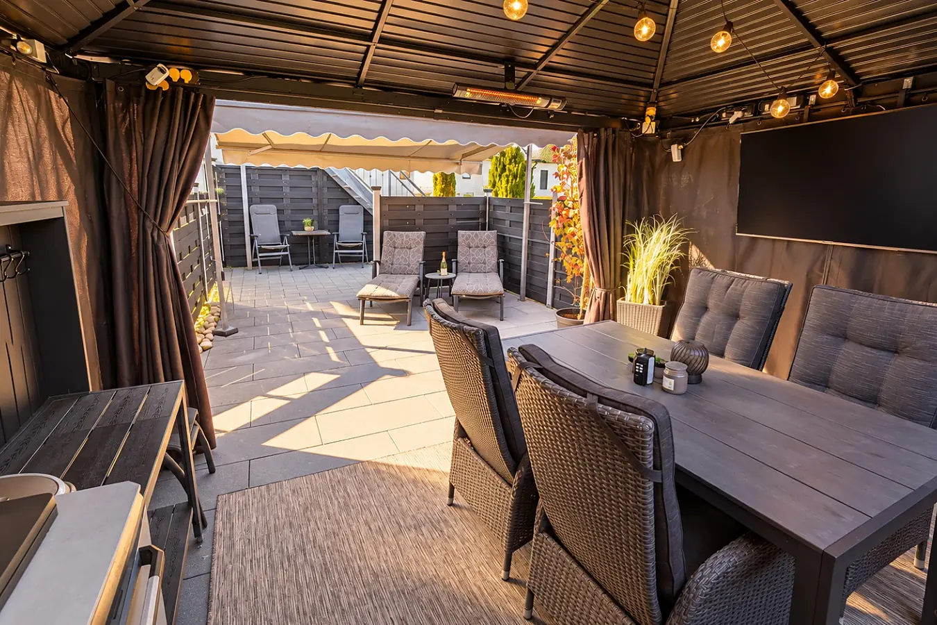 Blick aus dem Pavillon auf die Terrasse: großer Esstisch mit sechs Korbsesseln unter Lichterkette und Fernseher, seitlich Vorhänge, dahinter die offene Terrassenfläche mit Liegestühlen.