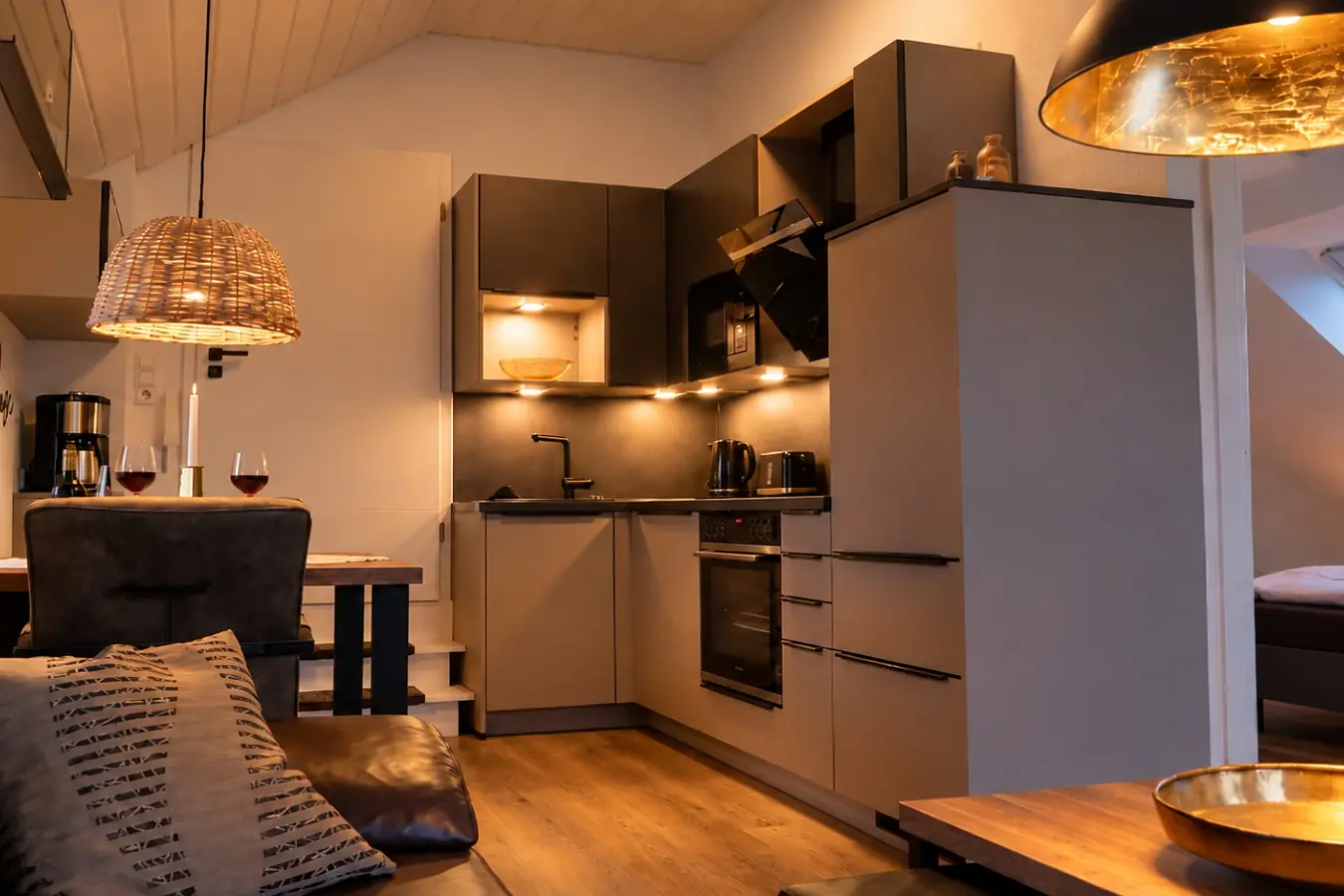 Blick von der Couch auf die Küche: graue Küchenzeile über Eck mit Backofen, Kochfeld, Dunstabzug, Wasserkocher und beleuchteten Hängeschränken, davor der Esstisch mit zwei Gläsern Rotwein unter einer Korbleuchte.