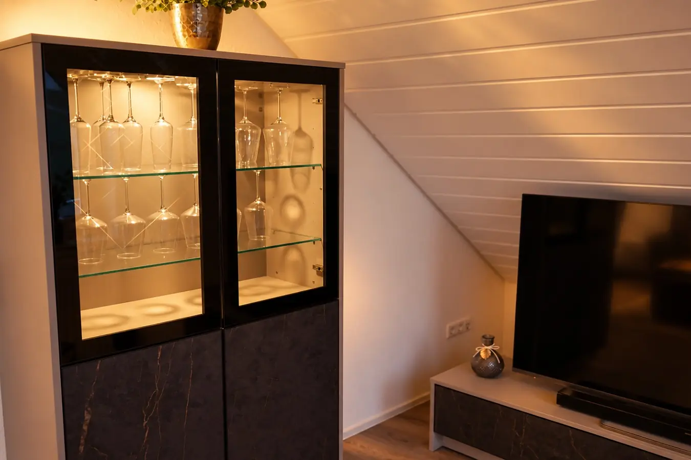 Beleuchtete Glasvitrine mit Wein- und Sektgläsern neben dem Fernseher auf einem Sideboard, darüber die weiße Dachschräge der Wohnung.