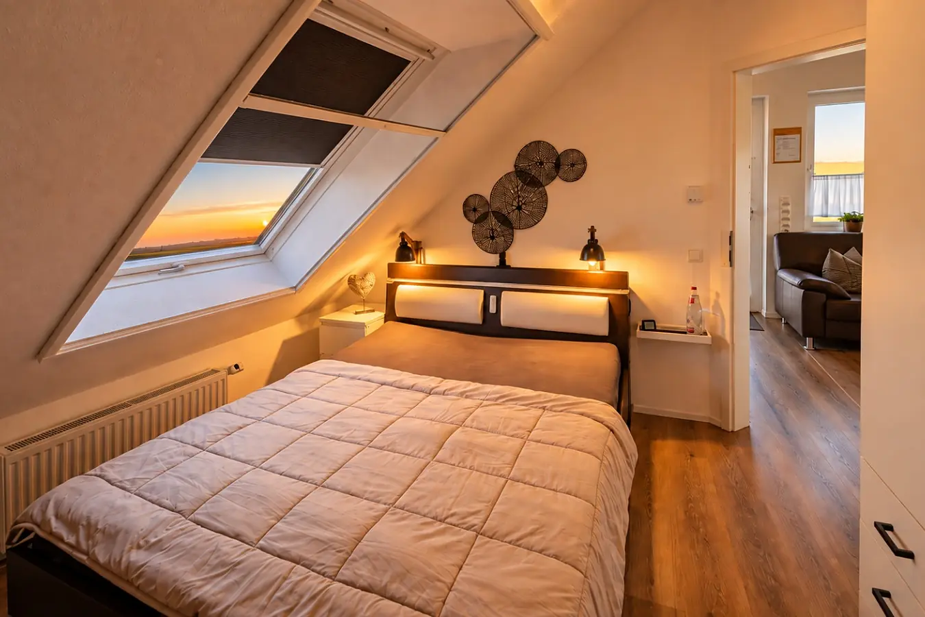 Schlafzimmer unter der Dachschräge: Doppelbett mit heller Tagesdecke, darüber ein Dachfenster mit Blick in den Abendhimmel, an der Wand Dekoscheiben aus Holz, rechts die offene Tür zum Wohnbereich.