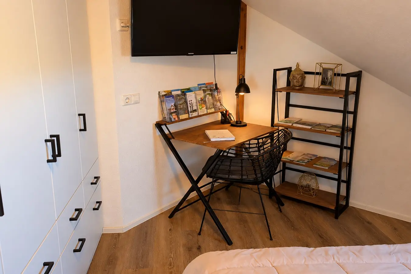 Schlafzimmer mit weißer Einbauschrankwand über die ganze Wand, daneben ein kleiner Schreibtisch mit Stuhl, Lampe und Prospekten, darüber ein Fernseher an der Wand, rechts die Bettkante.