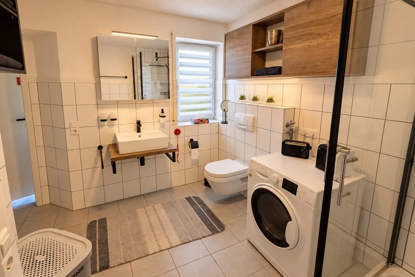 Bad mit weißen Fliesen: Waschbecken mit Spiegel unter einem Fenster, daneben das Wand-WC und die Waschmaschine, links die Dusche hinter Glas, auf dem Boden ein gestreifter Läufer.