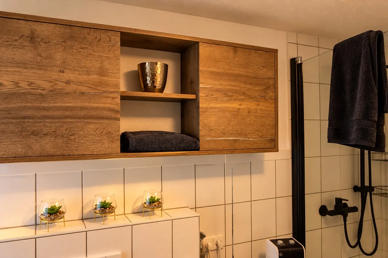 Duschbereich des Bads: schwarze Regendusche mit Handbrause an weißen Fliesen, daneben ein dunkles Handtuch, darüber ein Hängeschrank in Holzoptik mit offenem Fach und Deko.