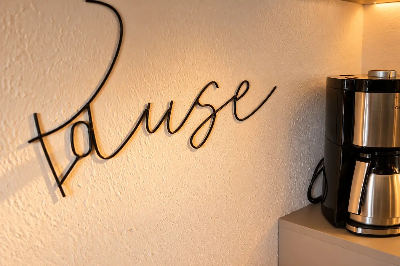 Schriftzug «Pause» aus schwarzem Metall an der cremefarbenen Wand, daneben eine Filterkaffeemaschine mit Thermoskanne auf der Arbeitsplatte.